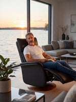 Homme assis dans une chaise inclinable noire TOCO Meubles en cuir, design moderne avec base en bois près d'un lac.