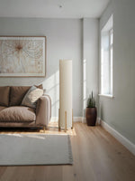Lampadaire cylindrique Luméa de TOCO Meubles avec abat-jour en tissu beige et pieds en bois clair dans un salon