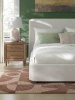 Grand lit Paloma TOCO Meubles en tissu bouclé blanc cassé avec tête de lit enveloppante, sur un tapis abstrait rose.