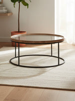Table de salon ronde Nora de TOCO Meubles avec plateau en verre et cadre en bois naturel sur pieds en métal noir
